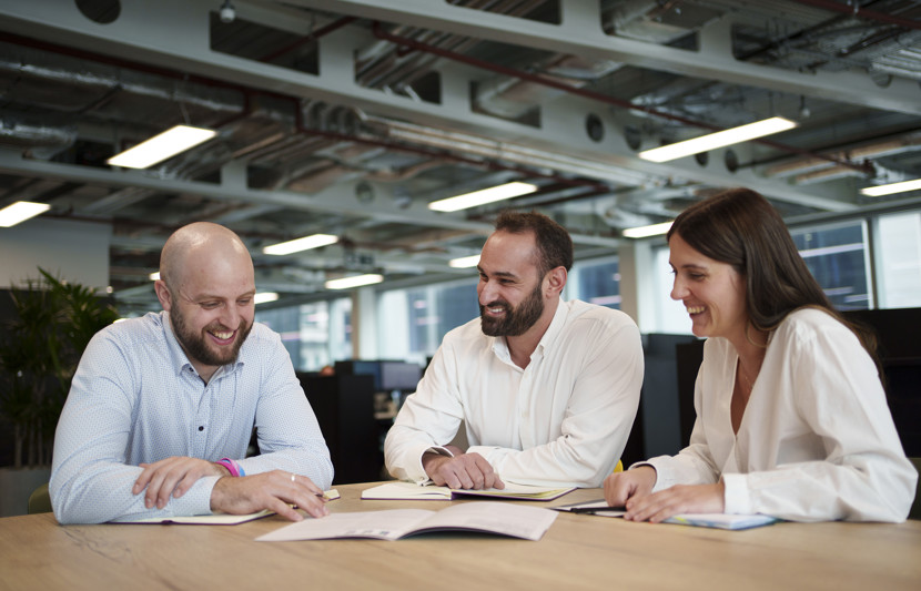 Three employees gathered round an informal meeting table with open plan office in background