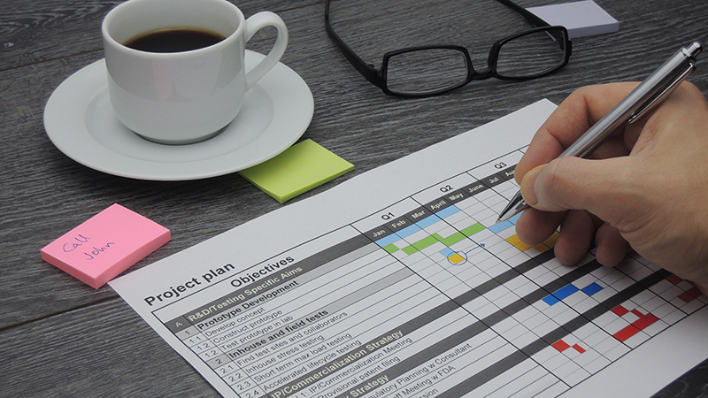 Close up on project manager's hand with pen as he reviews project plan. Other items on desk include reading glasses, white coffee cup, and post-it notes