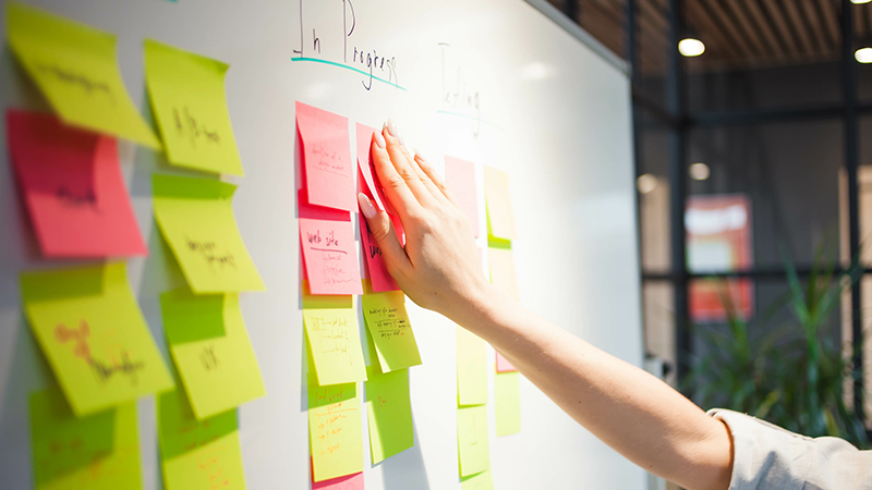 Close up on project manager's hand as she sticks post-it note to whiteboard while reviewing project progress against procedure. Note is among many bright yellow and pink notes arranged under progress headings.
