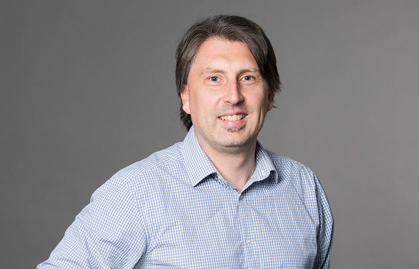 Daniel Coates RPS Technical Director - Survey and Registered Cadastral Surveyor smiles at the camera. Wearing blue check shirt, grey photo background.