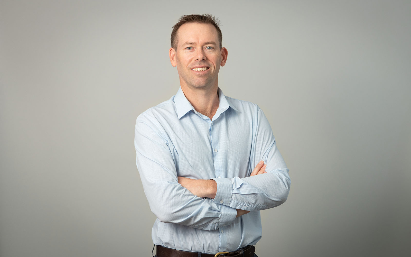 RPS Senior Principal Surveyor and Registered Surveyor, David Ada, smiles at the camera with crossed arms. Light blue shirt and grey background. 