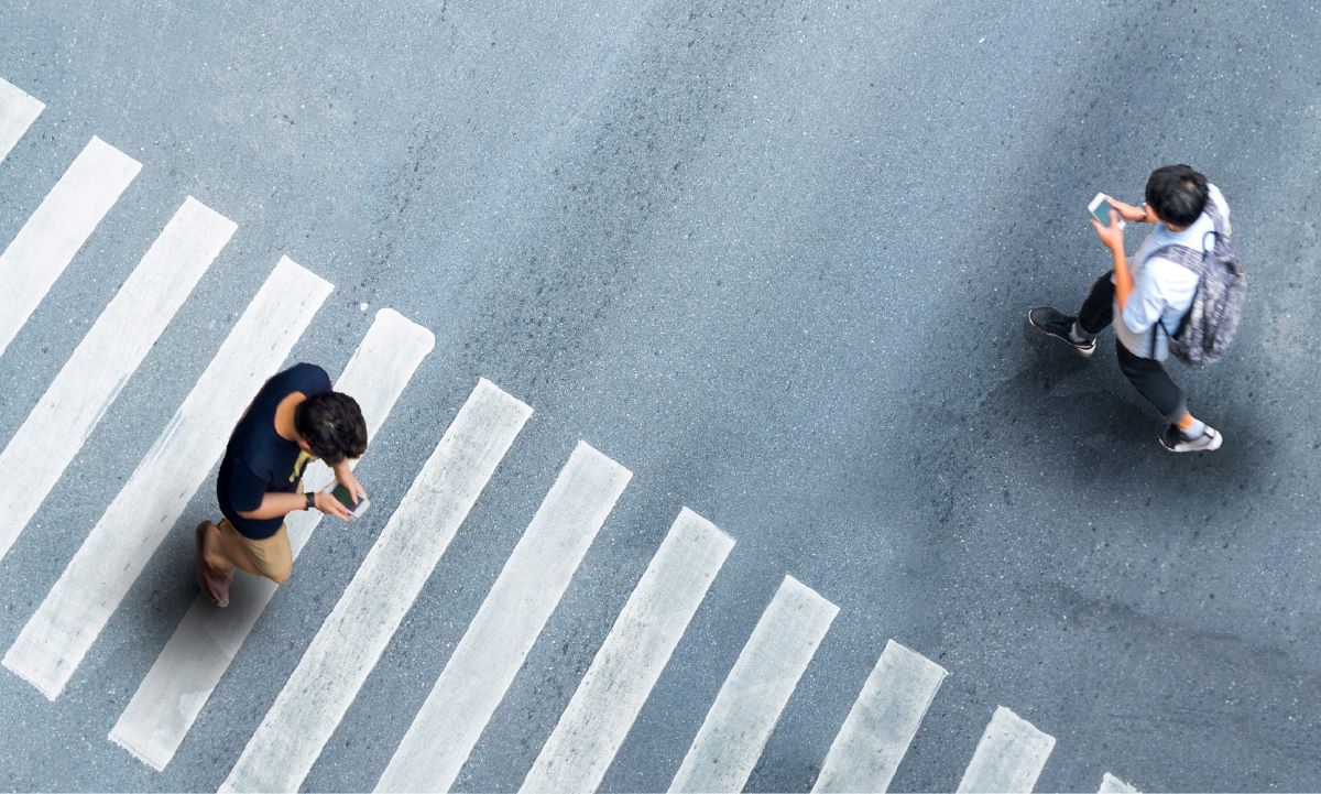 An overview shot of two men on opposite sides of a crosswalk and both are looking down at their phones as they walk 