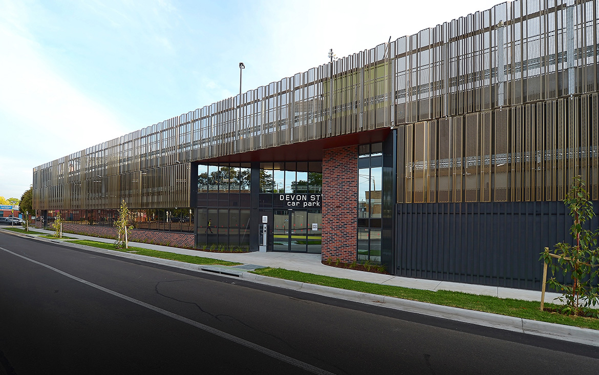 View of Devon Street carparking structure looking down the street along the facade on a sunny day. Bitumen road in the foreground, before grassed verge and footpath in front of building. Newly planted street-level trees and cloudy blue skies above.