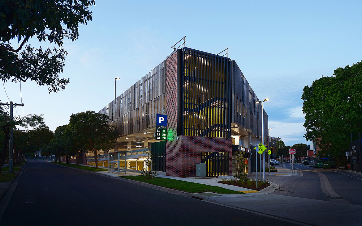 Twilight photograph of Devon Street carpark taken from the road showing lighting through metal facade to parking levels inside. 'P' Parking digital sign affixed to the building marks the entrance.
