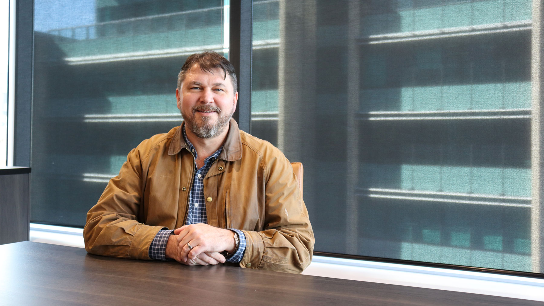Des Cloake, RPS Principal Landscape Architect, Brisbane Queensland, Australia.
Des sits at table and smiles at camera with hands crossed. Wearing brown jacket and blue plaid shirt.