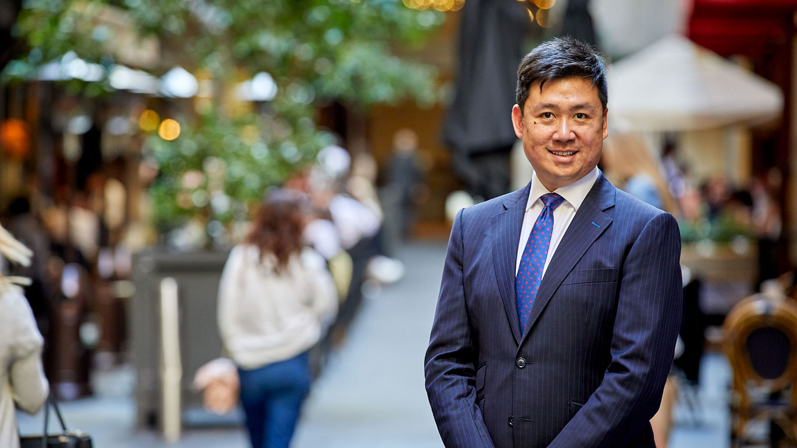 Dr Merrill Lee poses for photo in Sydney CBD. Wearing a blue suit and tie and is smiling at the camera. People in the background walking by are out of focus.