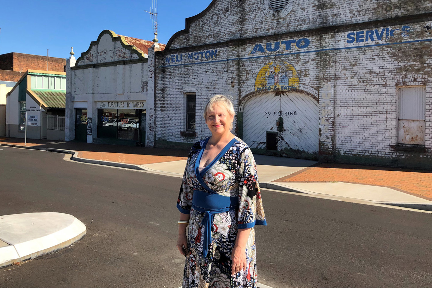 RPS Technical Director Engagement stands in country town street wearing a floral Japanese-inspired blue and red dress. Her backdrop is of a historical mechanic shop named Wellington Auto Services.