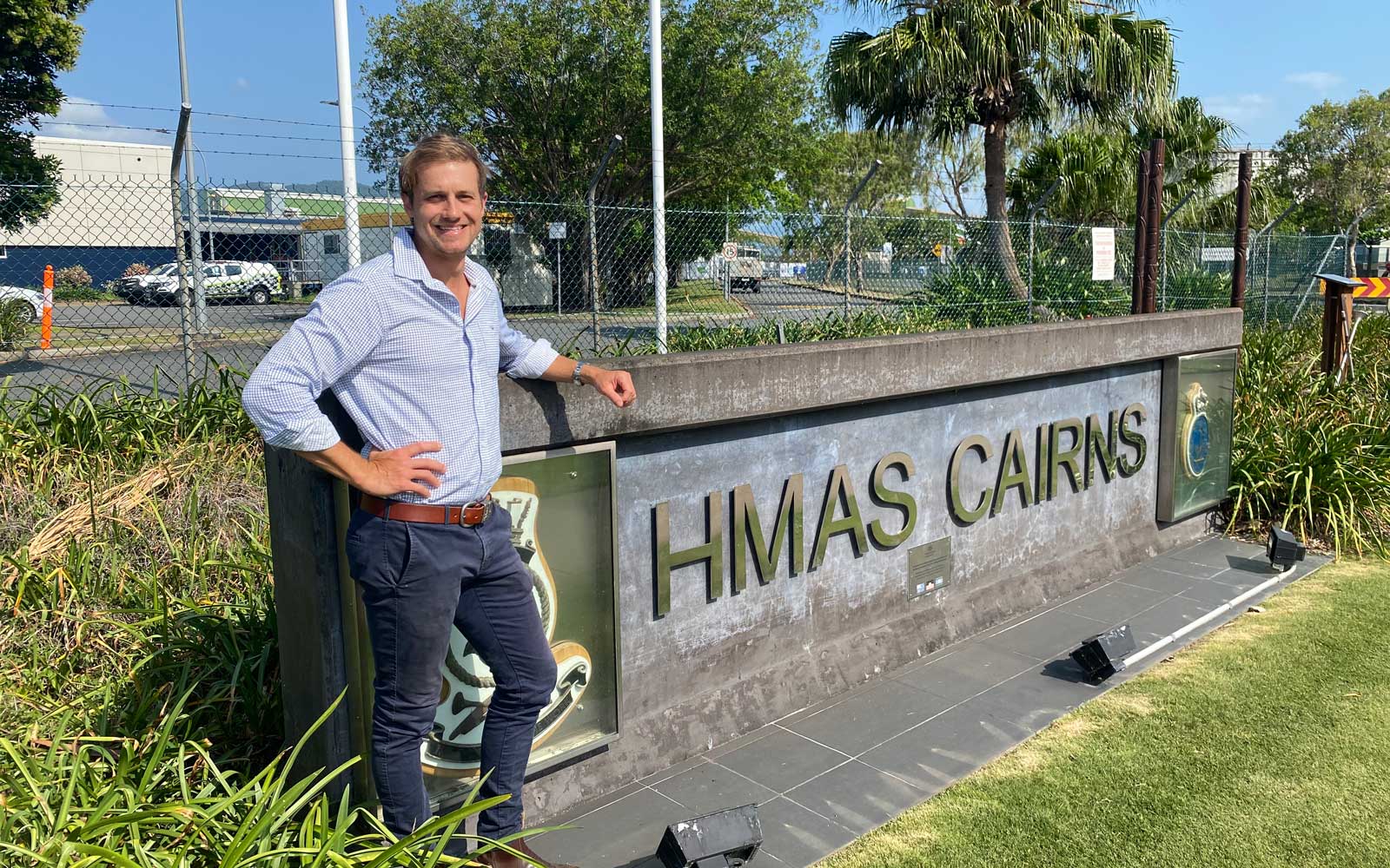 RPS Project Manager, Lee Leitner stands in front of a HMAS Cairns sign surrounded by grass and shrub. He wears a blue shirt and jeans and has his right arm in a pose. 