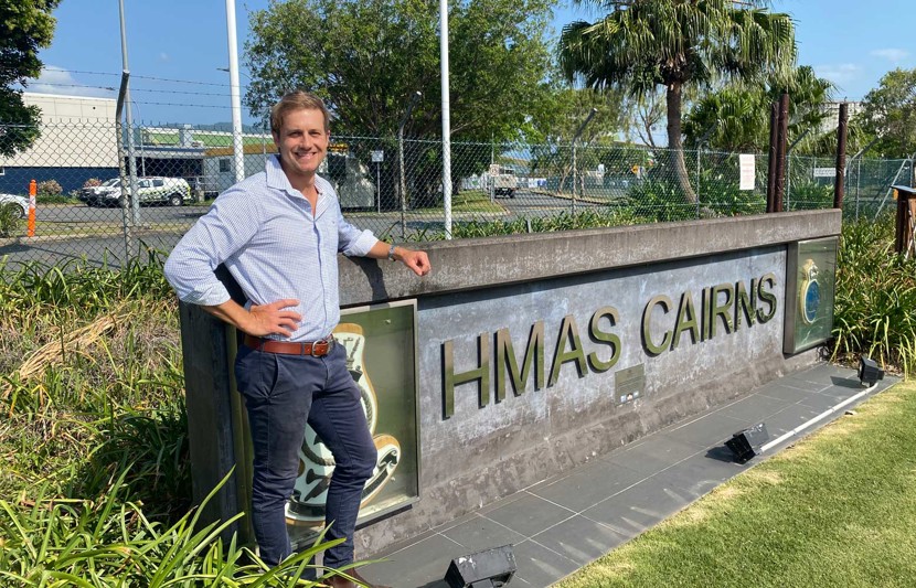 RPS Project Manager, Lee Leitner stands in front of a HMAS Cairns sign surrounded by grass and shrub. He wears a blue shirt and jeans and has his right arm in a pose.