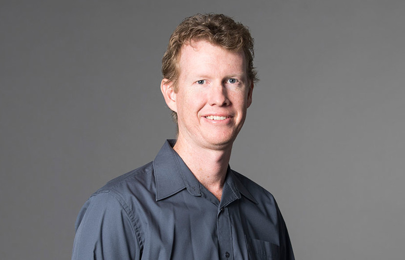 Simon Sager, RPS Principal Surveyor smiles at the camera wearing a grey button up shirt
