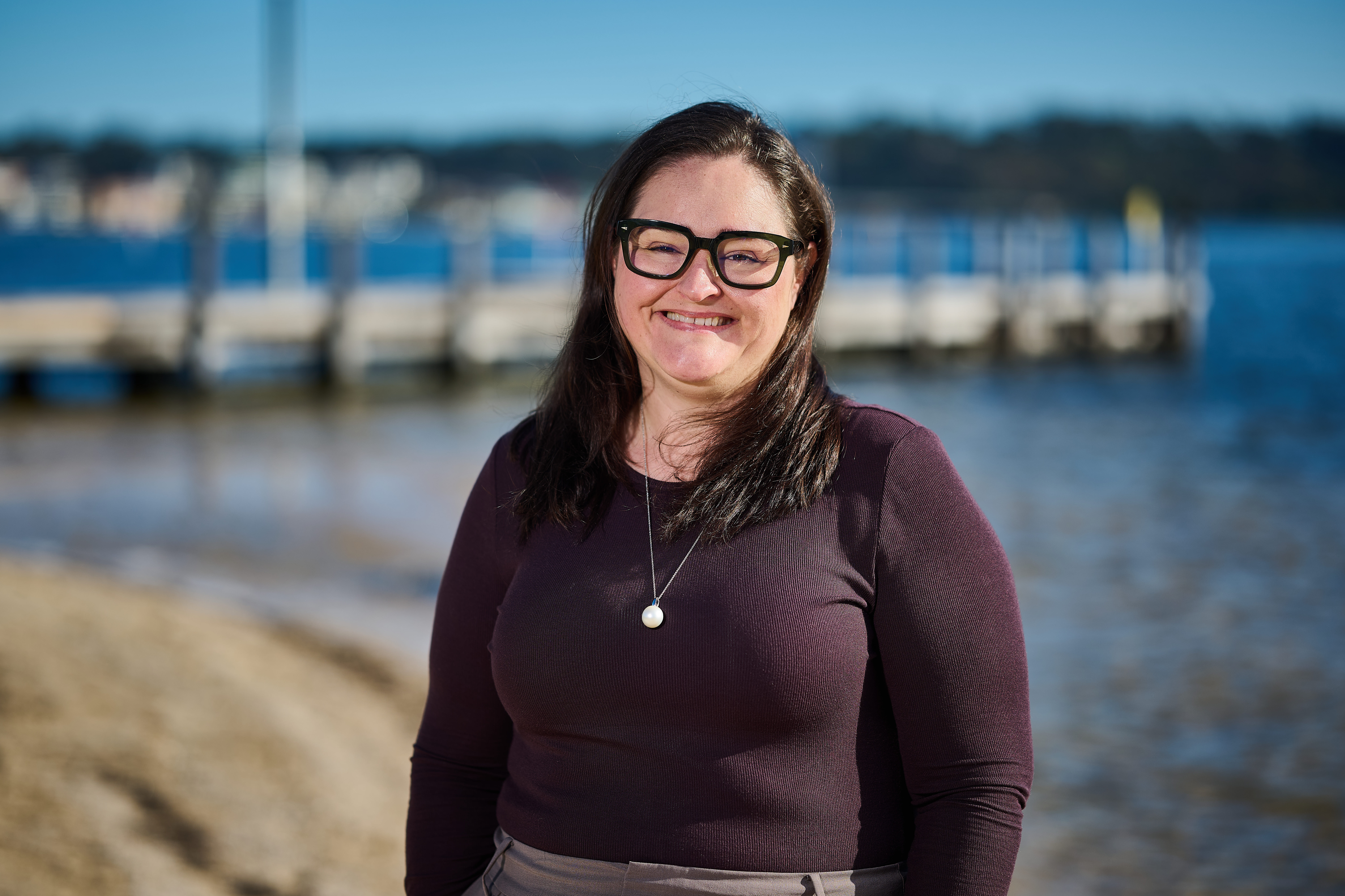 RPS' Senior Oceanographer Emma Foster smiles into camera with a beach and jetty as her backdrop. 