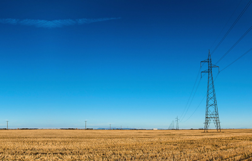 Transmission lines extend into the distance across rural farmland in regional Australia. Blue clear skies and recently harvested cropped pasture in the foreground.