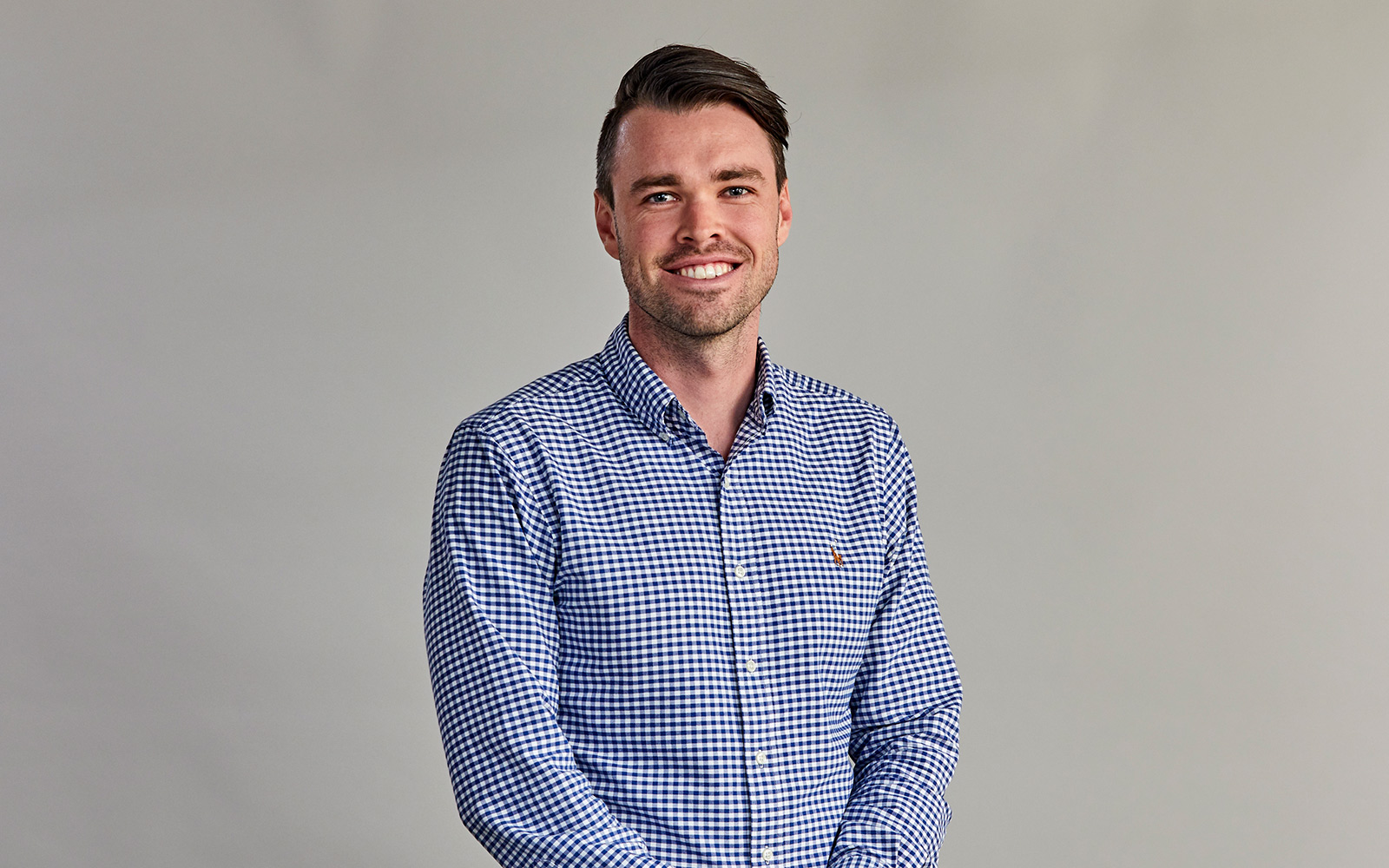 RPS Principal Surveyor, Luke Sharkey smiles at the camera. Blue check shirt, grey background.
