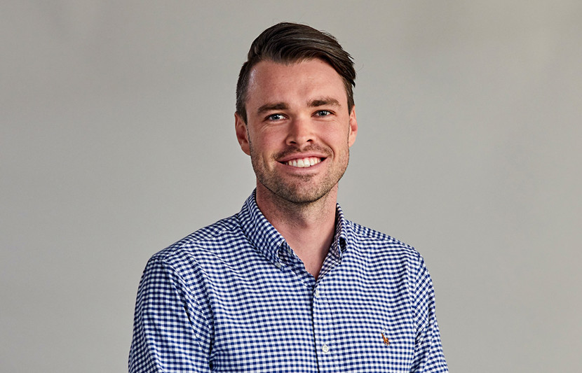 RPS Principal Surveyor, Luke Sharkey smiles at the camera. Blue check shirt, grey background.